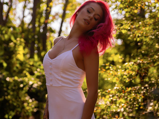 KaylieRosse, con i capelli rosa, canotta bianca e parte del petto coperta da bottoni, in piedi alla luce del sole, con gli occhi chiusi, la testa reclinata all'indietro e le mani ai lati, mostra il sedere e i piccoli seni..