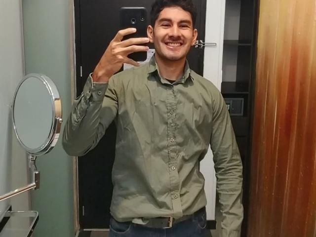 KevinAndres, sorridente, che tiene un telefono, indossa una camicia verde a bottoni, jeans blu, è in piedi in un bagno, riflesso nello specchio, specchio rotondo a sinistra, porta di legno a destra..