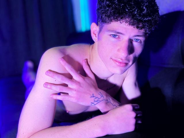 MatiaAgt sin camisa, con cabello negro rizado, ojos azules y tatuajes en la mano derecha, inclinado hacia adelante con una ligera sonrisa, con las manos cerca del pecho, en un ambiente iluminado con luz púrpura..
