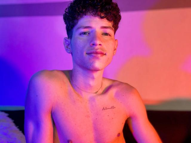 MatiaAgt, torse nu, avec des cheveux bruns bouclés, des yeux verts et un petit tatouage sur la poitrine, portant un collier, sous un éclairage rose et bleu. MatiaAgt a un léger sourire..