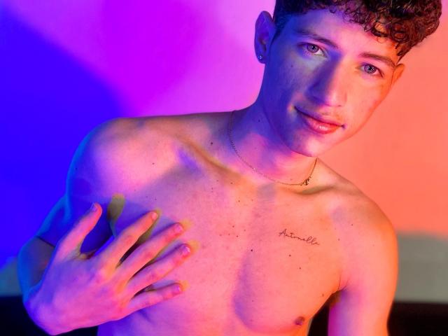 MatiaAgt, avec des cheveux bruns bouclés, torse nu, une main sur la poitrine, un tatouage avec l'inscription "acquellc" sur le côté droit, un éclairage violet et orange, un léger sourire, un collier, des yeux bleus et un visage mignon..