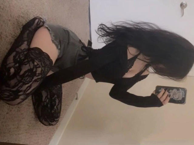 GermanVixen-hot on knees, wearing black lace thigh-highs, gray shorts, and a black top, exposing her ass and cleavage, taking a mirror selfie with a phone.
