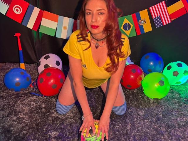 QueenScarletSecret está arrodillada sobre una alfombra gris, vistiendo una camisa amarilla, calcetines azules a rayas y un collar, rodeada de pelotas de colores y un bate, con un banderín detrás..