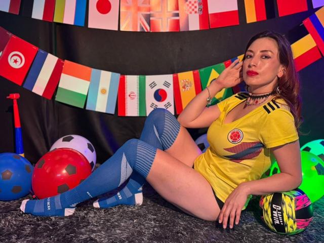 QueenScarletSecret avec une grande fesse, de petites seins, vêtue d'un maillot de foot jaune, de chaussettes bleues, avec du rouge à lèvres, un collier noir, assise sur un tapis gris, avec des ballons de foot, des drapeaux et un sac de ballons de foot..
