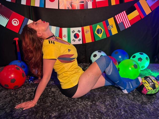 QueenScarletSecret avec des cheveux roux, un haut de sport jaune, un short noir, des chaussettes bleues jusqu'aux genoux et un collier, penchée en arrière sur un tapis gris, entourée de ballons de foot et de drapeaux colorés..