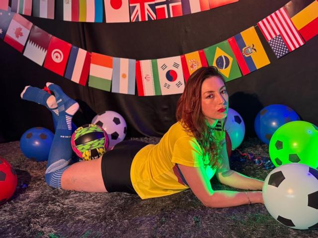 QueenScarletSecret, acostada boca abajo, vestida con una camisa amarilla, pantalones cortos negros, calcetines azules hasta la rodilla y calcetines a rayas, con balones de fútbol y globos de colores a su alrededor, sobre una alfombra..