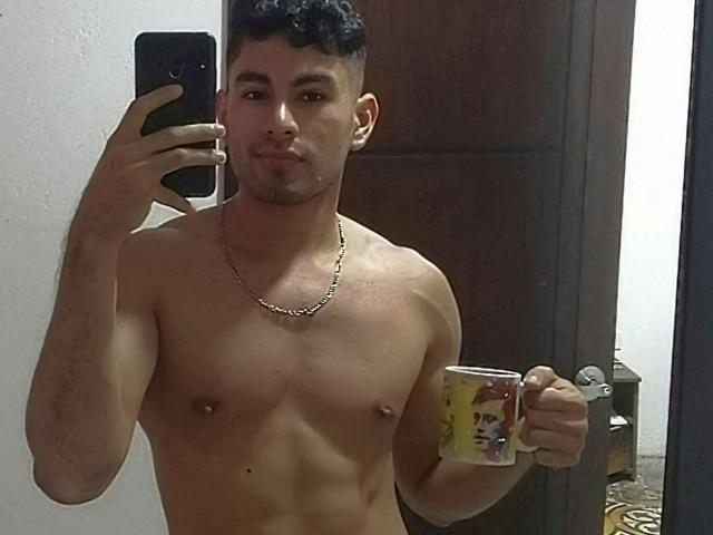 KevinAndres, torse nu et musclé, tenant une tasse de café dans sa main droite, prend un selfie devant un miroir avec sa main gauche. Il porte un collier en chaîne et a des cheveux noirs et bouclés..