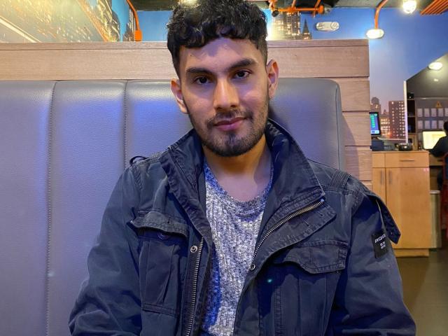 KevinAndres, un hombre con cabello rizado y barba, que lleva una chaqueta vaquera sobre una camisa gris con estampado, sentado en una cabina gris, sonriendo ligeramente..