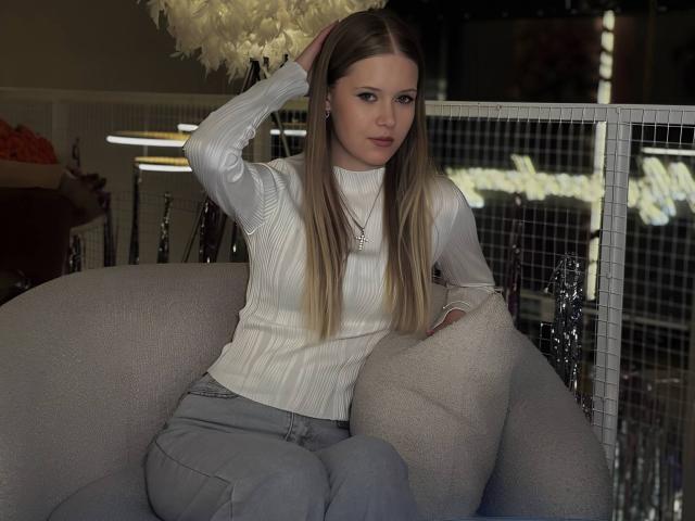 Lilliana Whang tem cabelos loiros longos, veste um suéter branco texturizado, calças cinzas e um colar. Ela está sentada em uma cadeira cinza e macia, com uma mão no cabelo..