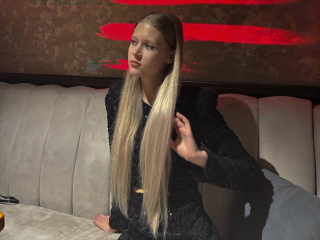 Lilliana Whang mit langen blonden Haaren, die ein schwarzes, flauschiges Kleid trägt, sitzt in einer grauen Kabine, blickt leicht zur Seite und lächelt dabei, ihre Hand ist in der Nähe ihres Gesichts..