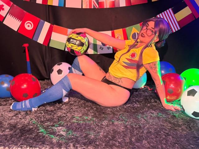 LilithDark69 est assise sur un tapis gris, vêtue d'un maillot de foot jaune, d'une culotte noire, de chaussettes bleues et de lunettes, tenant un ballon de foot, avec des ballons colorés et une batte à côté..