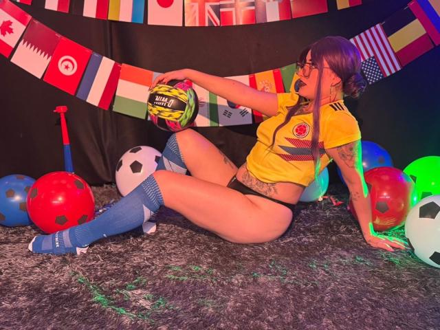 LilithDark69 mit Tattoos, einem gelben Fußballtrikot, blauen Socken und schwarzen Unterhosen, hält einen Fußball und ist umgeben von bunten Bällen und Flaggen auf einem flauschigen Teppich..