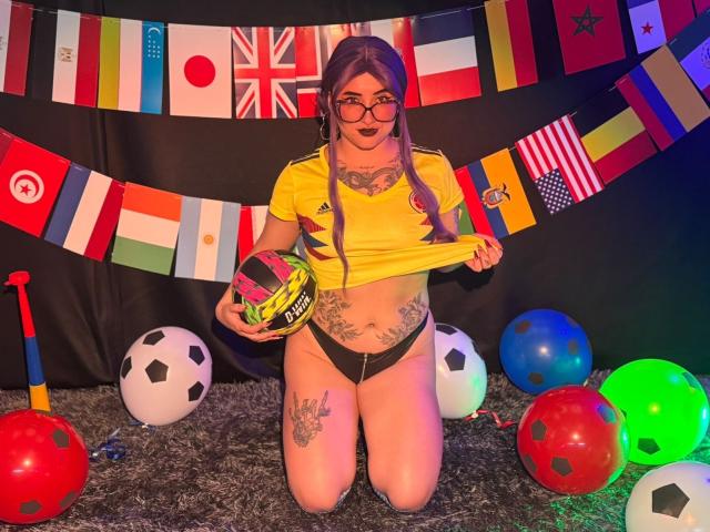 LilithDark69 ajoelha-se em frente a uma decoração com tema de futebol, vestindo uma camisa de futebol amarela levantada para mostrar seus seios, calcinhas pretas, e segurando um capacete, cercada por balões coloridos..
