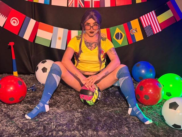 LilithDark69, avec ses cheveux violets et ses lunettes, portant un haut de sport jaune, des chaussettes bleues jusqu'aux genoux, et tenant un casque rose, est assise les jambes écartées, entourée de ballons de foot colorés..
