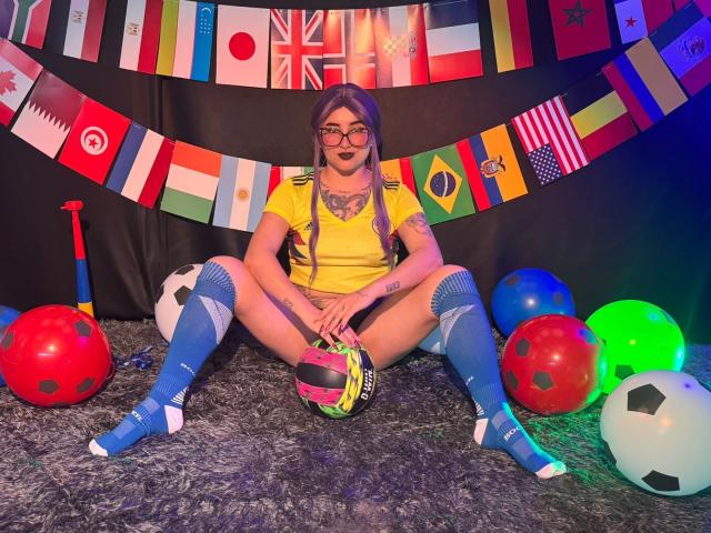 LilithDark69, avec des lunettes, un t-shirt jaune, des chaussettes bleues jusqu'aux genoux, assise les jambes écartées, tenant un ballon de foot, entourée de ballons colorés et de ballons de foot, avec du rouge à lèvres noir et un tatouage sur la poitrine..