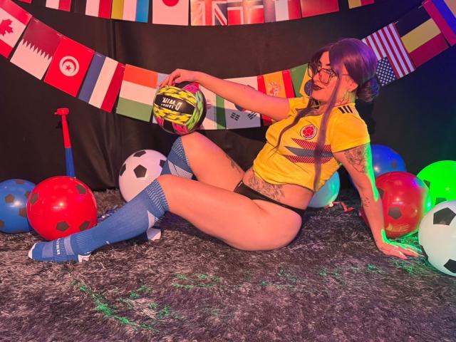 LilithDark69 con cabello morado, gafas, camiseta amarilla de fútbol, ropa interior negra, calcetines azules hasta las rodillas, sosteniendo un balón de fútbol, sentada sobre una alfombra gris, rodeada de balones de fútbol y banderas..