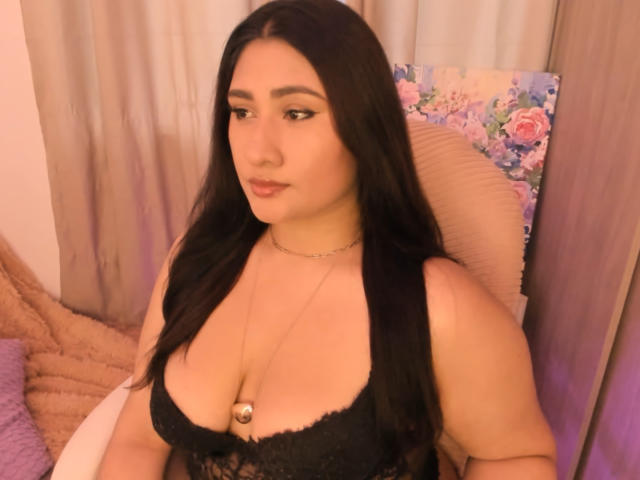ElyRoss with long black hair, medium skin, and full tits in black lace lingerie, wearing a necklace with a pendant, sitting in a pink, floral, and beige room.