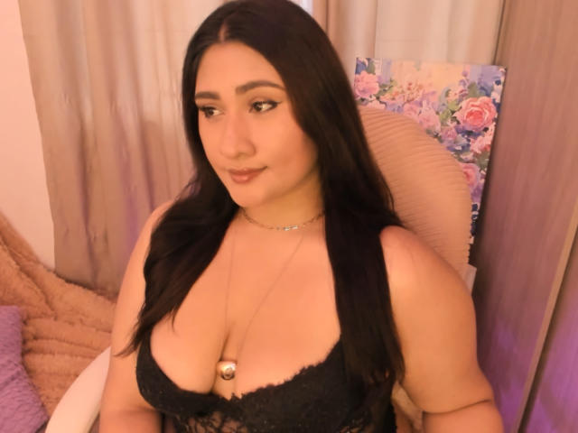 ElyRoss has dark, wavy hair, full eyebrows, and is wearing black lace lingerie that shows her big, round tits. She's looking down with a slight smile.