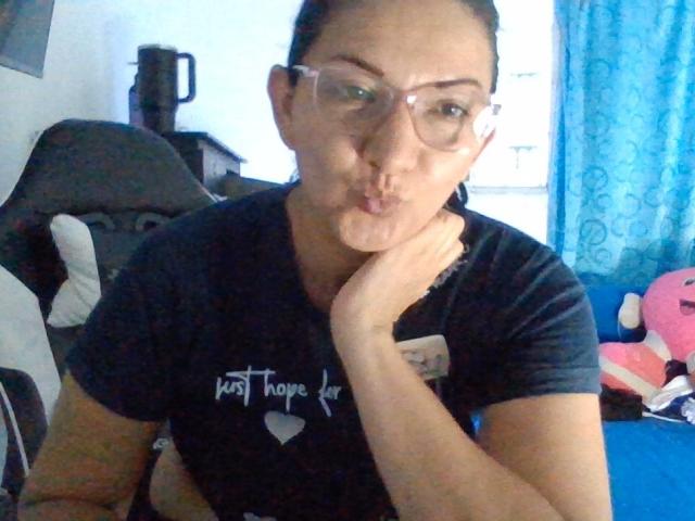 AngelBlue69, met volle lippen, een bril op, en een zwart T-shirt, leunt met haar kin op haar hand en heeft een licht geïrriteerde of fronsende uitdrukking. Haar bruine haar is naar achteren gebonden..