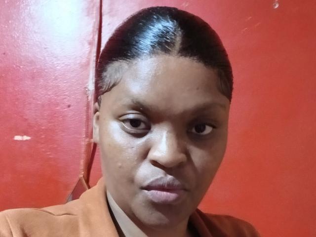 NickiAli with dark skin, slicked-back hair, small pimple on cheek, serious expression, wearing brown shirt, against red wall. Her face looks slightly tired.