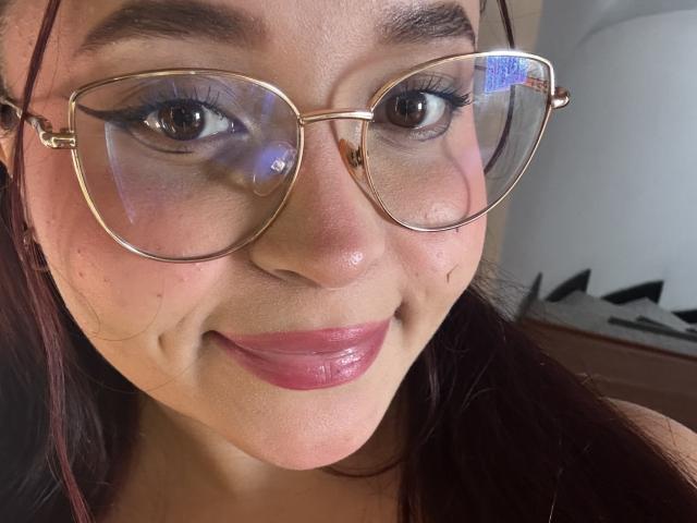 SamyMancini with shiny pink lips, wearing gold-rimmed glasses, has dark brown eyes and long dark hair, smiling at the camera. Her skin is light with a few freckles.