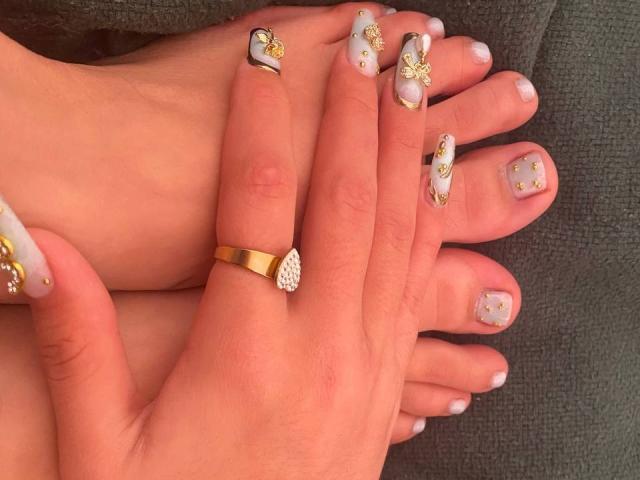 SweetSolei69's hands with manicured nails featuring white, gold, and black designs, including small gold flowers and gems. She wears a gold ring with a diamond on her index finger.