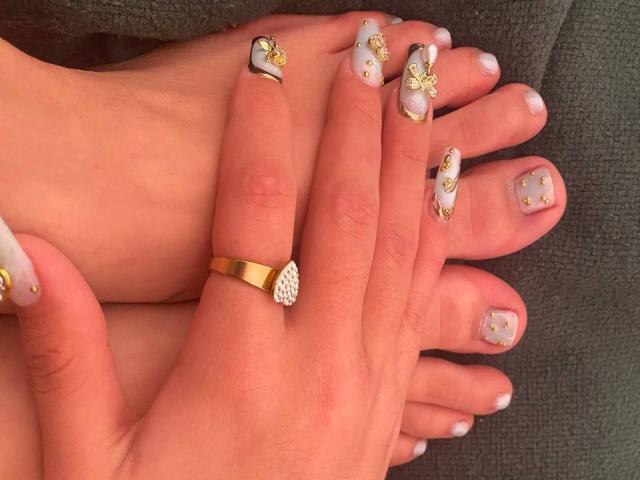 SweetSolei69's hands and feet, showing manicured nails with white, gold, and silver designs, wearing a gold ring with a diamond. Her toes are painted similarly.