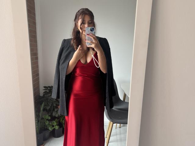 LinaFoxyHot in a red satin dress, black jacket, holding phone, mirror selfie, brown wavy hair, medium tits, fair skin, white wall, black chair, potted plant, brick.