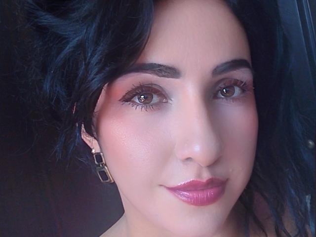 Il viso di LuluFrancaise, con i capelli scuri e ondulati, gli occhi marroni, il rossetto rosa e le labbra rosa lucide, indossa orecchini quadrati e guarda direttamente l'obiettivo..