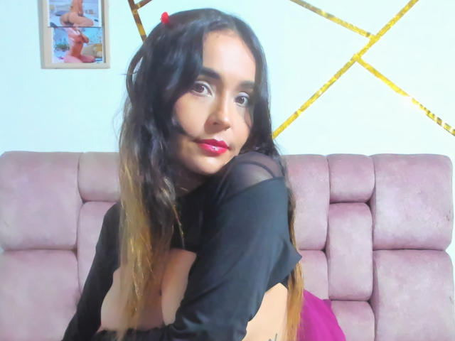 MindyLove with long black hair, red lipstick, and black top, showing her tits, leaning on a pink sofa, smiling, and wearing a small red devil horn on her head.