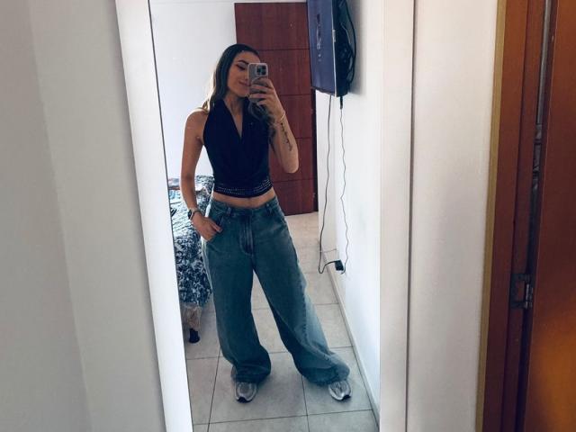 AdharaCooperr with long dark hair, black sleeveless top, and blue baggy pants, standing in front of mirror, holding phone, tattoos visible on right arm, white wall behind.