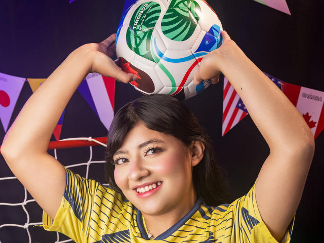 NekoPam con i capelli scuri, sorridente, che tiene una palla da calcio sopra la testa, indossa una maglia da calcio gialla e blu, con le mani alzate. I suoi seni sono parzialmente visibili..