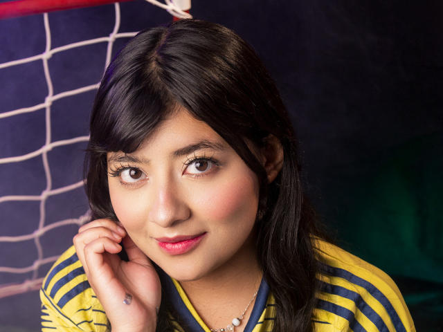 A NekoPam tem cabelo preto e liso, olhos castanhos, batom rosa e um sorriso discreto. Ela está vestindo uma camisa listrada em amarelo e azul com um colar branco pequeno..