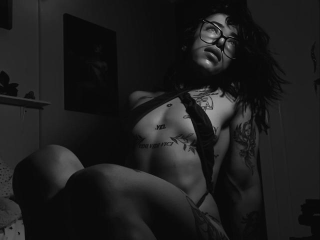YeiSela, usando óculos, com cabelo escuro e tatuagens, vestindo um sutiã de renda preto, sentada com os joelhos levantados, com os seios à mostra, olhando para cima com a boca ligeiramente aberta, em uma iluminação escura e melancólica..