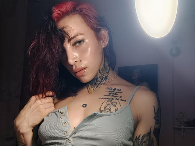 YeiSela met een glanzende huid, rood en zwart haar, die een grijze blouse draagt, met meerdere tatoeages, haar blouse vasthoudend, kijkend naar de camera met een licht geopende mond..