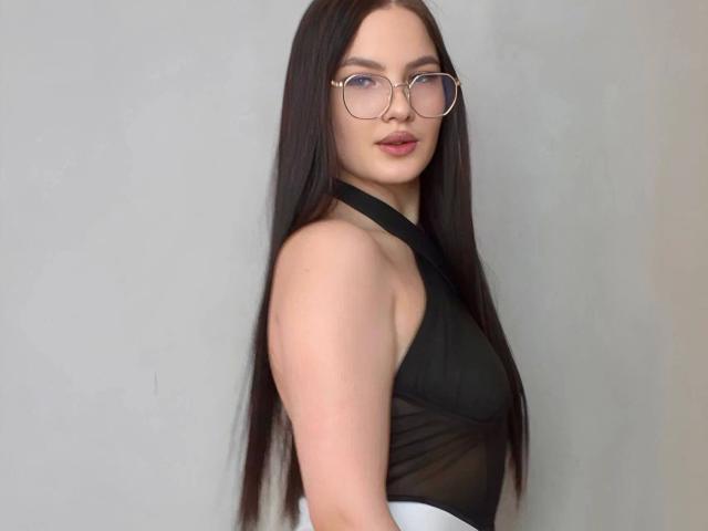 MilanaBeautty-hot has long dark brown hair, wears glasses, and a black halter top, showing her bare shoulders and small, perky tits. She's looking over her shoulder.
