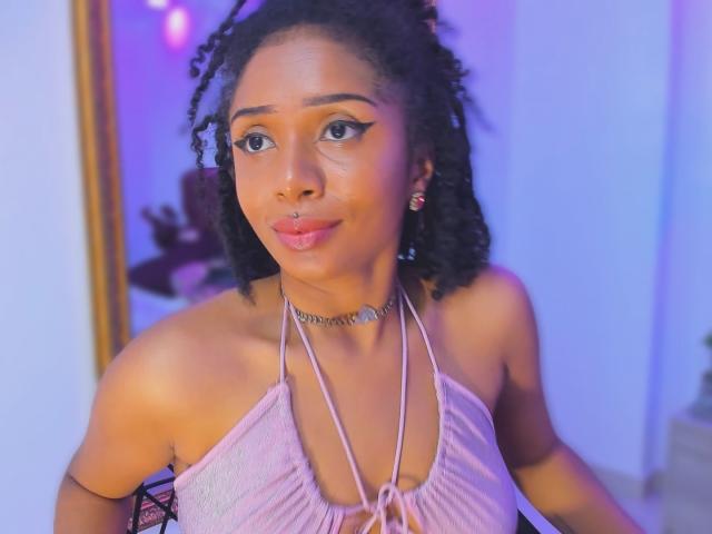 MeganWlliams with dark curly hair, light brown skin, small nose ring, pink lip gloss, pink halter top, choker, looking at camera, blue and purple lighting.