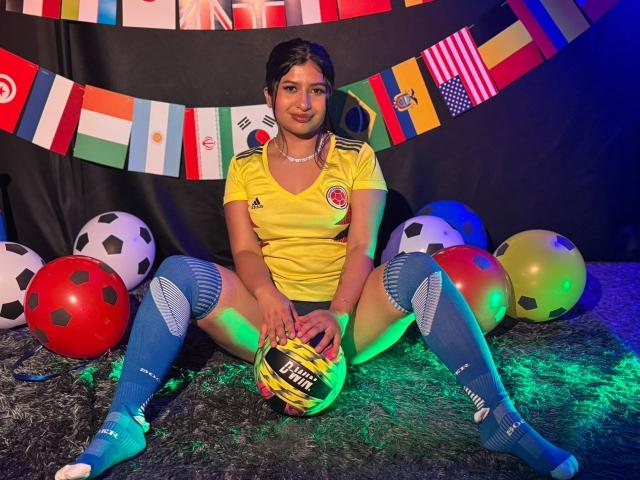 ZarayWhitMoreLove, com a parte de trás da calça de futebol à mostra, usando uma camisa amarela, meias azuis até o joelho, segurando uma bola, cercada por bolas de futebol, bandeiras coloridas, sentada em um tapete verde, cabelo escuro, colar, sorrindo..