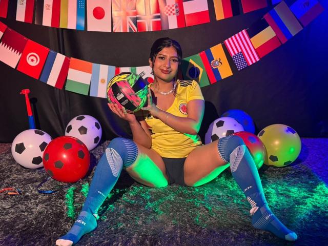 ZarayWhitMoreLove, sitting with legs spread, wearing a yellow soccer jersey, blue knee socks, and black panties, holding a soccer ball, surrounded by colorful balloons and soccer balls.