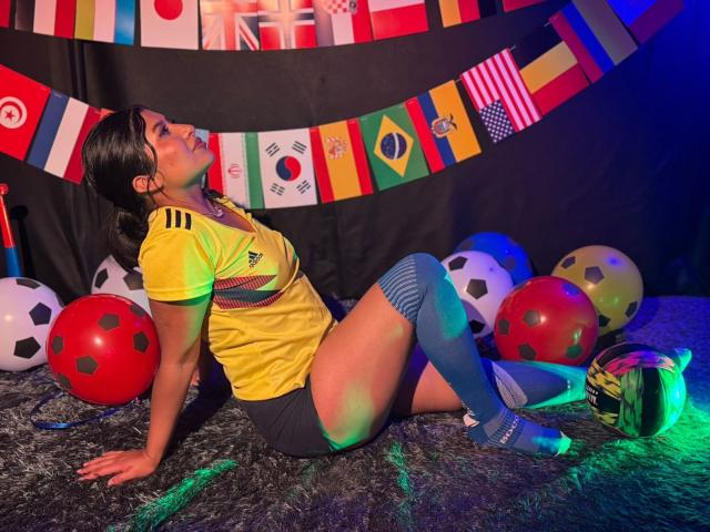 ZarayWhitMoreLove, Asian woman with black hair, yellow soccer shirt, blue knee socks, and black shorts, sitting on a furry black rug, surrounded by soccer balls and flags.