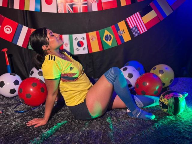 ZarayWhitMoreLove, sentada con la espalda apoyada en el suelo, vestida con una camiseta de fútbol amarilla, calcetines azules y pantalones cortos grises, rodeada de balones de fútbol, banderas coloridas y luces azul verdosas..