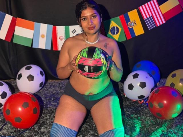 ZarayWhitMoreLove, oben ohne mit grauen Unterhosen, hält einen bunten Fußball, umgeben von Fußbällen, trägt blaue Kniestrümpfe, auf einem grauen, strukturierten Boden, mit bunten Flaggen im Hintergrund..