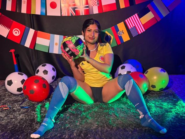 ZarayWhitMoreLove, con un seno di media grandezza, indossa una maglia da calcio gialla, tiene una palla da calcio, calze blu fino al ginocchio, seduta su un tappeto grigio con palle da calcio e bandierine colorate..