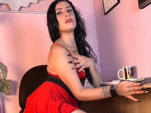 DemiMistral, avec de longs cheveux noirs, une robe rouge et des ongles noirs, est assise à un bureau, une main sur sa poitrine, regardant la caméra, montrant ses tatouages, tenant une tasse et portant une montre..