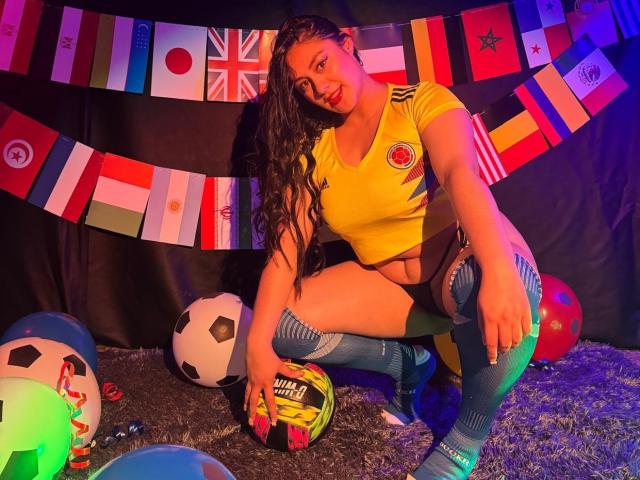 CristalZafiro met lang, krullend bruin haar, een geel voetbalsok, blauwe kniekousen en zwarte korte broek, hurkt over voetballen, met vlaggen op de achtergrond, kleurrijke lichten en een tapijt..