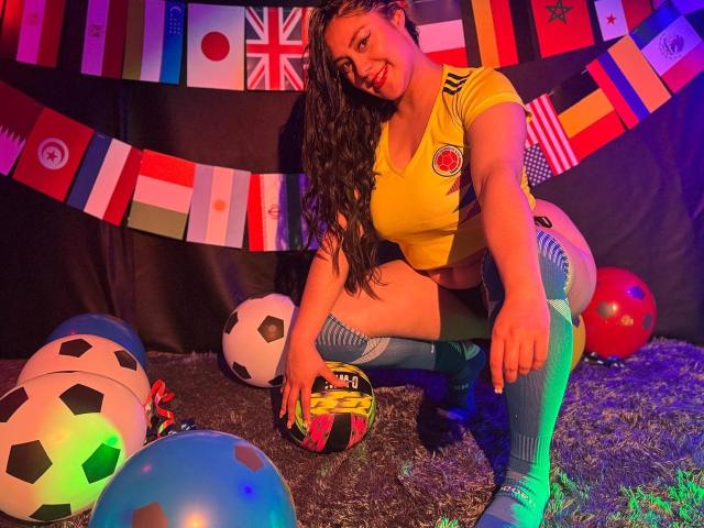 CristalZafiro mit langen, lockigen, braunen Haaren, gelbem Fußballtrikot, gestreiften Kniestrümpfen, in einer hockenden Position, lächelnd, einen Fußball haltend, umgeben von bunten Flaggen, Fußballbällen, auf einem grasbewachsenen Boden..
