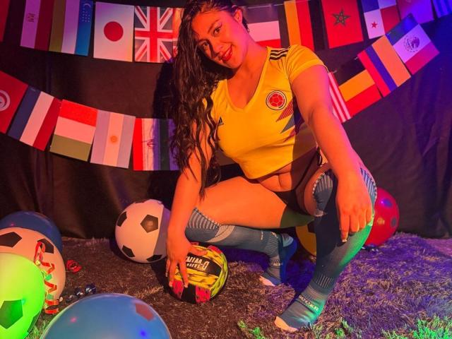 CristalZafiro squatting, wearing yellow soccer top, black shorts, striped socks, holding soccer ball, smiling, long curly hair, colorful flags, neon lights, soccer balls on gray carpet.