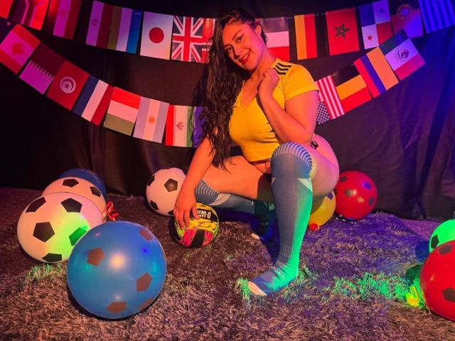 CristalZafiro met lang zwart haar, een gele top, blauwe kniekousen en gestreepte shorts, hurkt op een pluizig tapijt, omringd door voetballen en kleurrijke vlaggen..