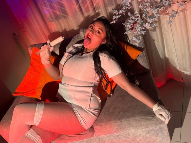 CristalZafiro with long black hair, tongue out, holding a popsicle, wearing a tight white dress, sheer stockings, white gloves, sitting on a couch, with orange pillows.