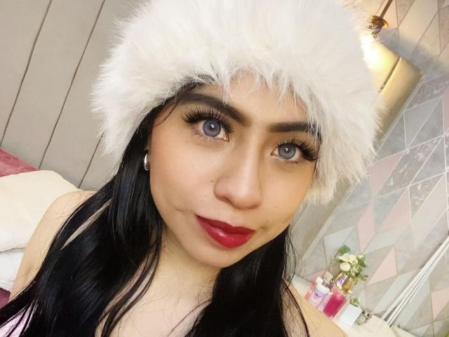 ZoeRedds with black hair, blue eyes, and red lipstick, wearing a fluffy white hat, looking into the camera, showing off her tits in a pink top.